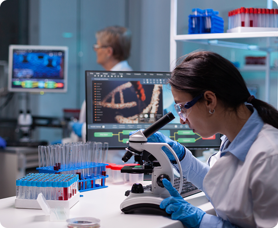Scientist examining samples under a microscope in a laboratory with computer screens and test tubes.