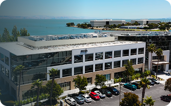 Modern office building with parked cars, palm trees, and a waterfront in the background.