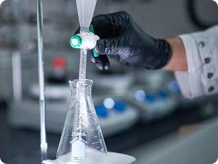 Gloved hand pouring liquid into an Erlenmeyer flask in a laboratory.