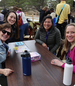 Four women smiling around a wooden table outdoors with water bottles and board games.
