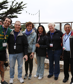 Six people smiling outdoors, standing together on a paved area with trees in the background.