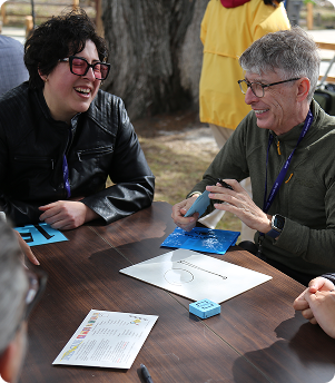 Two people laughing and playing a game at an outdoor table.