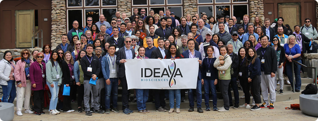 Large group of people posing outside a building, holding a banner that says "IDEAA."